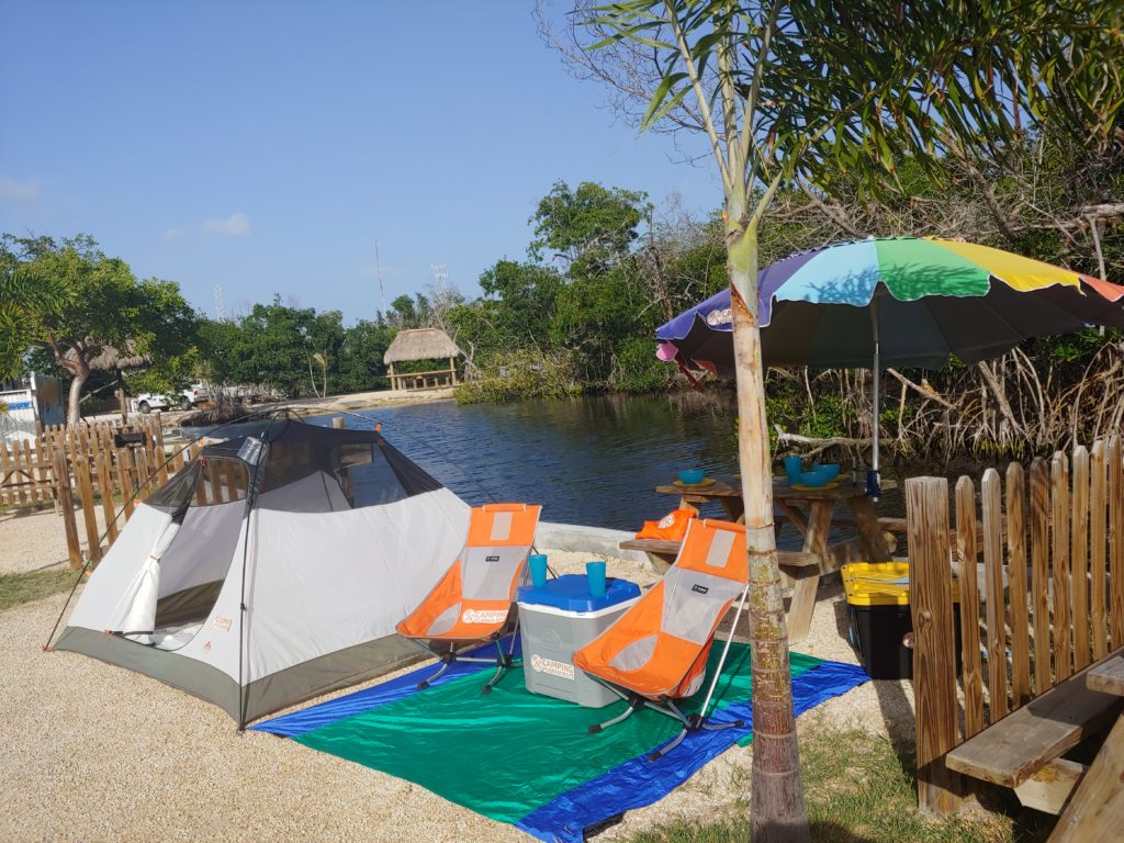 Kelty 4 person tent set up on a canal with 2 orange helinox beach chairs and a beach blanket underneath with a picnic table behind the chairs with an umbrella clamped to the side of the table. This is set on the banks of a mangrove lined canal in Florida at Leo's Campground.